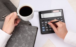Zwei Hände über einem Schreibtisch mit Unterlagen. Die Linke Hand hälte eine Weisse Kaffeetasse mit Kaffee. Die Rechte Hand tippt mit einem Stift auf einem Taschenrechner.